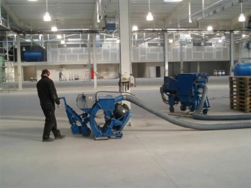 Osoba s ochrannými sluchátky obsluhující modrý podlahový stroj v průmyslové hale.