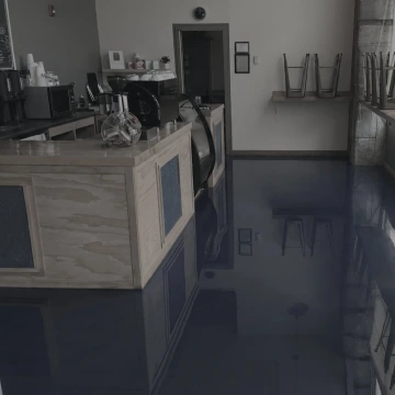 Kavárna s modrou zalitou podlahou, prázdné židle na stolech, pult s kávovarem a nádobami.